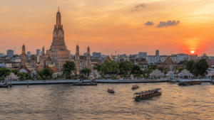 Wat Arun beside the Chao Phraya River with boats and the Bangkok skyline at sunset