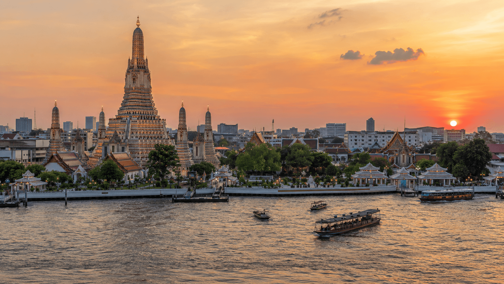 Wat Arun beside the Chao Phraya River with boats and the Bangkok skyline at sunset