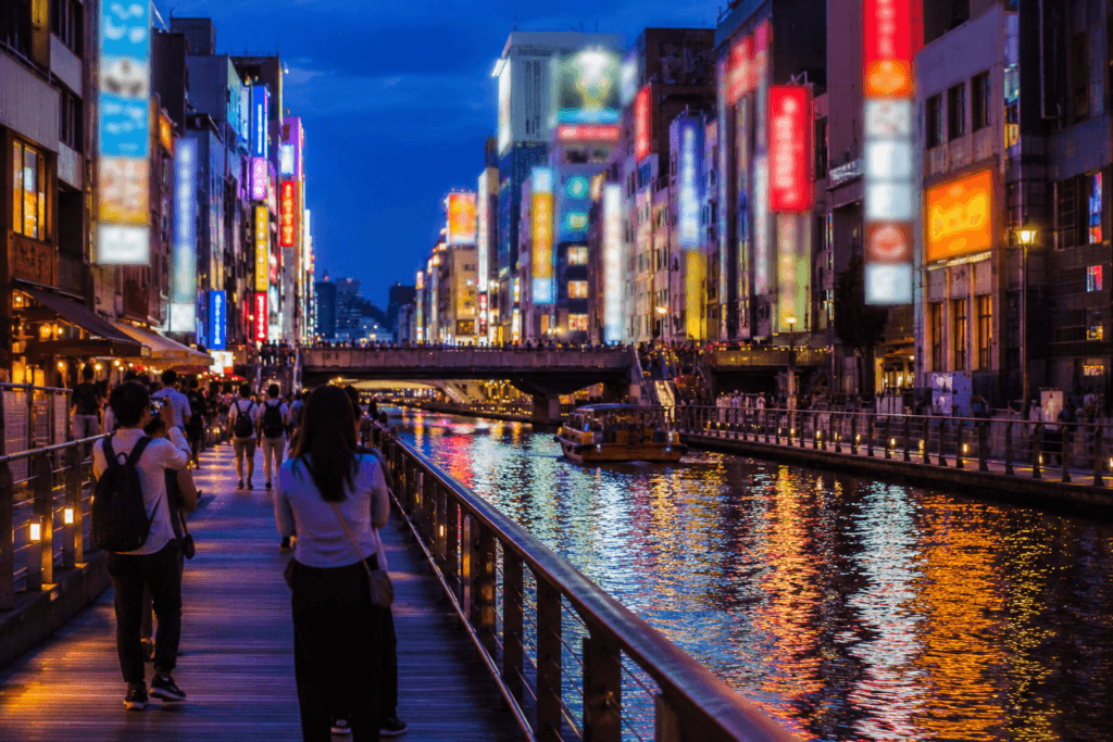 Travelers walking along a Dotonbori-style canal in Osaka at blue hour with neon lights reflecting on the water