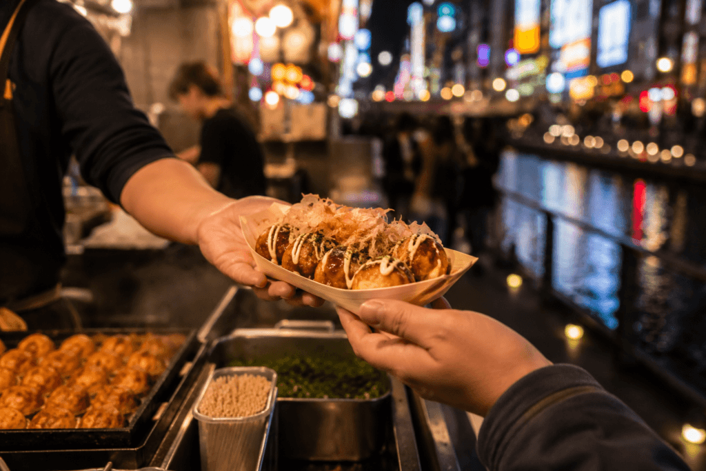 Fresh takoyaki being served at night with blurred neon lights and Dotonbori-style canal atmosphere in the background