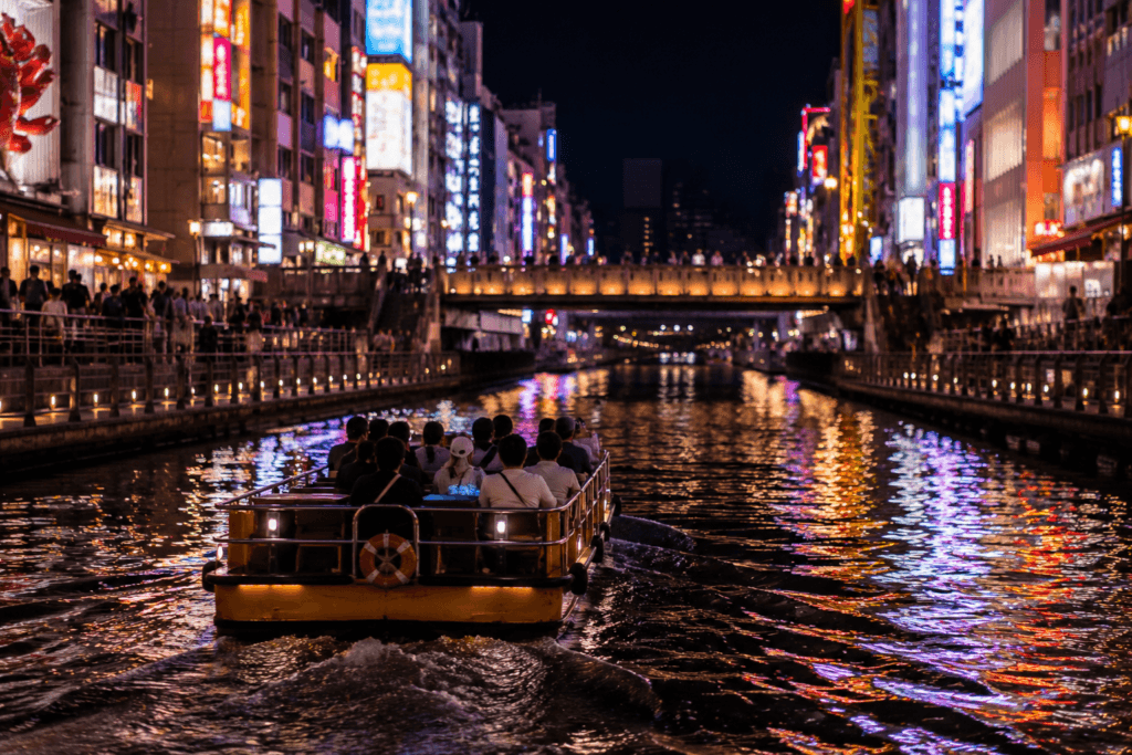 Small sightseeing boat cruising along a Dotonbori-style canal at night with neon reflections and crowded riverside walkways